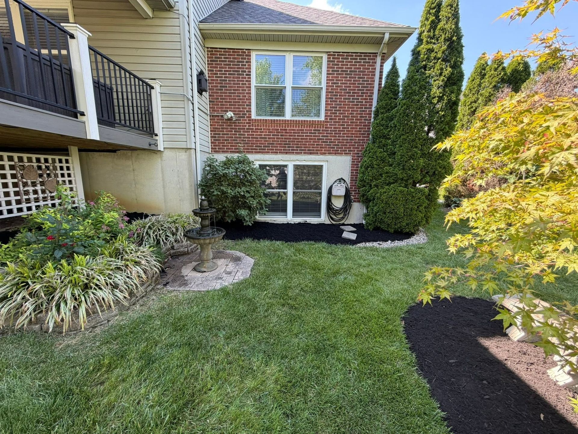 Small landscaped front yard with grass, shrubs, stone border, and a brick house exterior with windows.