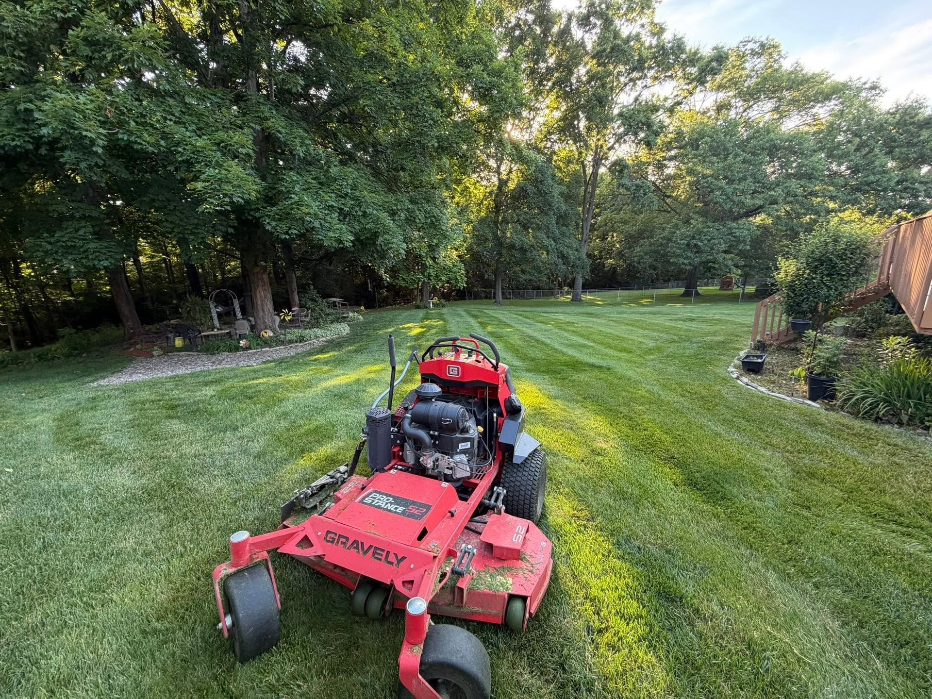 Red zero-turn mower on a striped grassy lawn beside trees and a wooden fence.