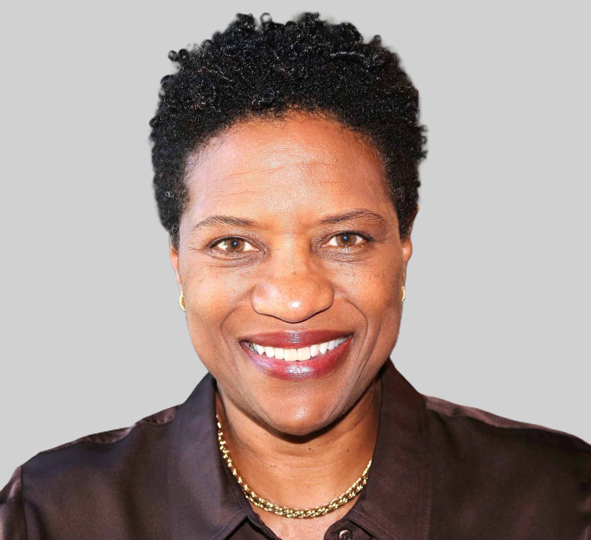 Portrait of a smiling person with short curly hair wearing a dark blouse and gold necklace on a light background