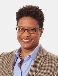Smiling person wearing glasses, blue shirt, and tan blazer against a white background