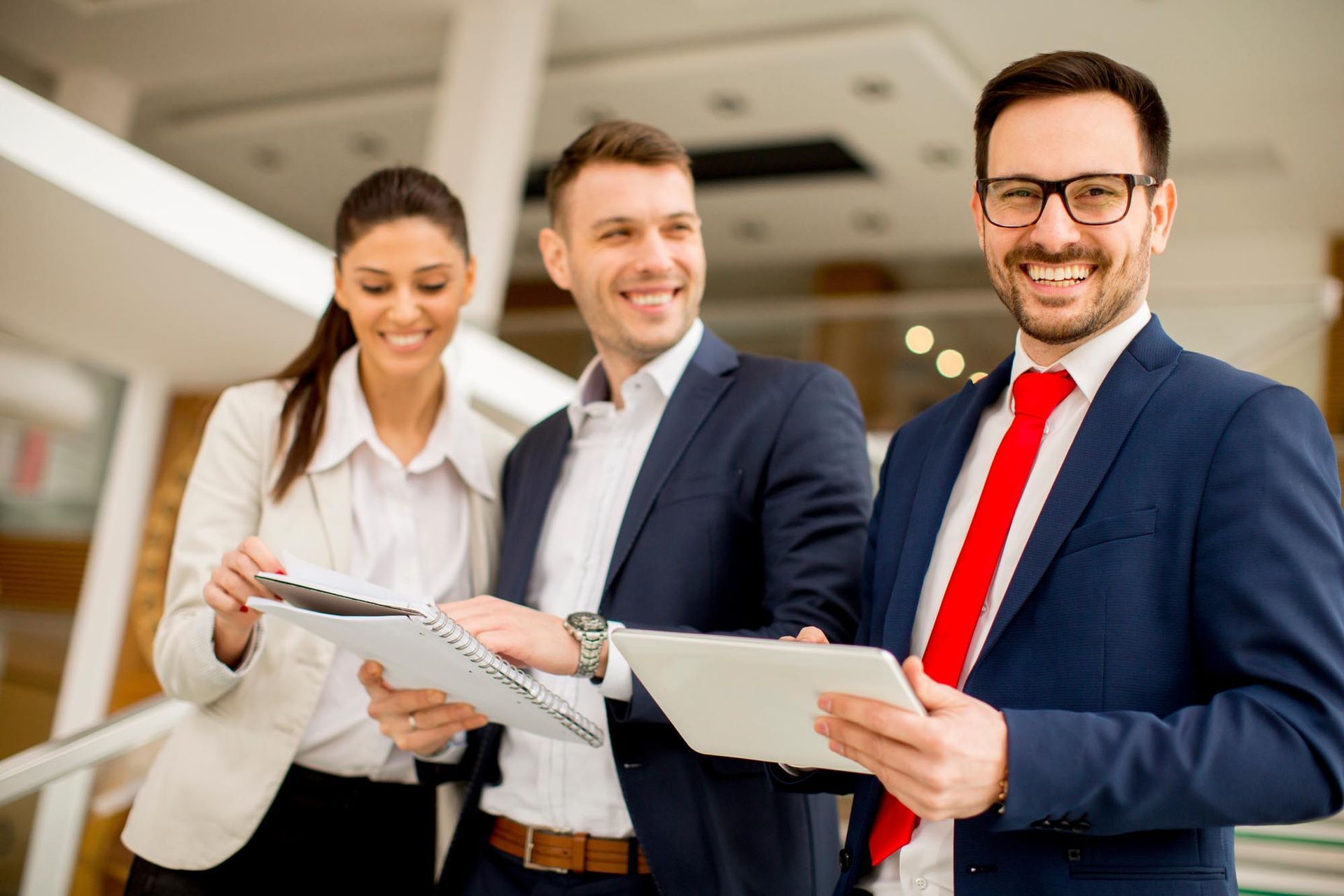 Three professionals in business attire stand in an office lobby, smiling as they review documents on a tablet and paper.