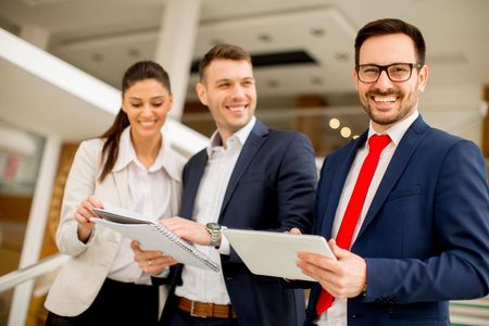Three business professionals in suits smile while reviewing documents and a digital tablet in a bright office space.