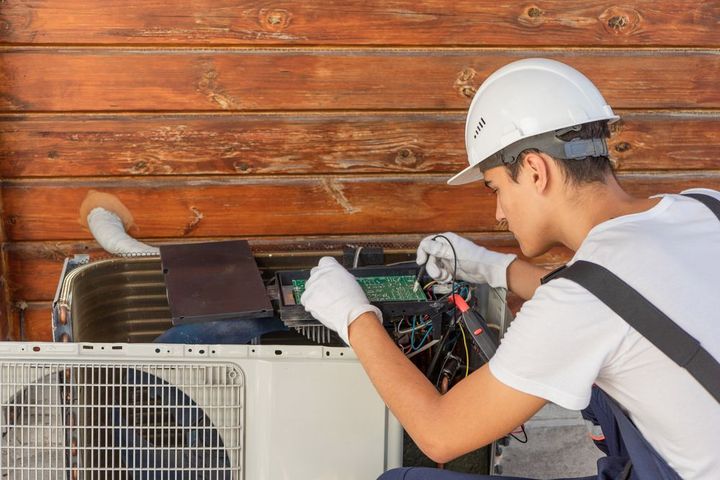 Un technicien portant un casque de chantier inspecte un climatiseur extérieur à l'aide d'outils, près d'un mur en bois.