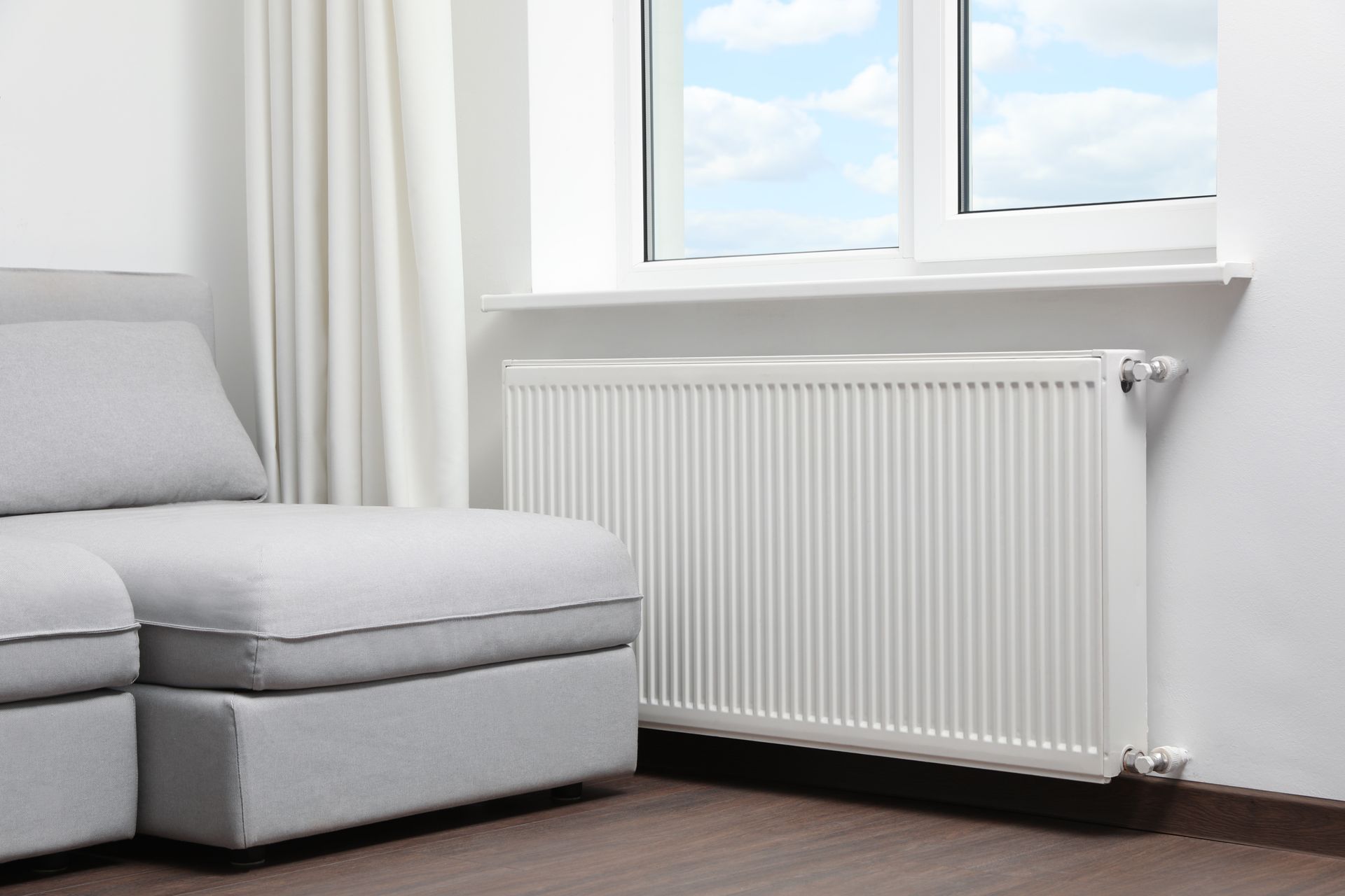 Coin salon minimaliste avec un canapé gris, un radiateur blanc et une fenêtre avec des rideaux transparents.