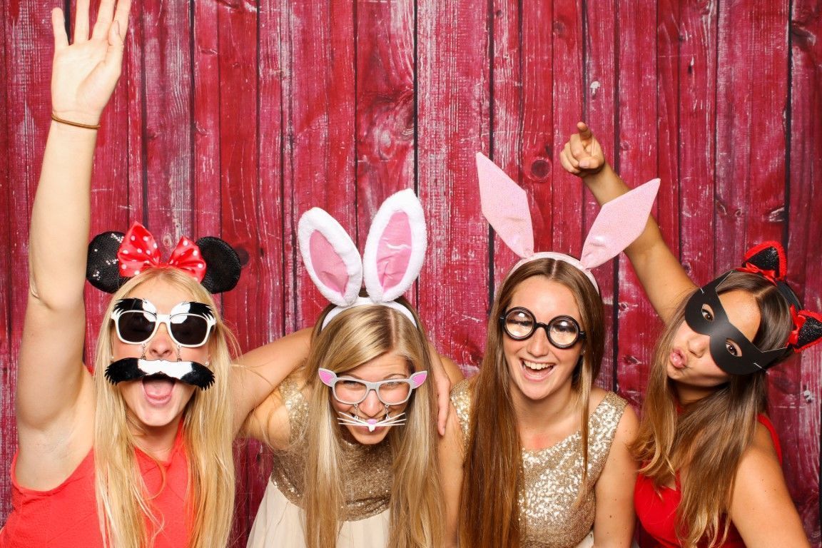 Four friends in playful costume accessories posing and smiling in front of a red wooden backdrop.