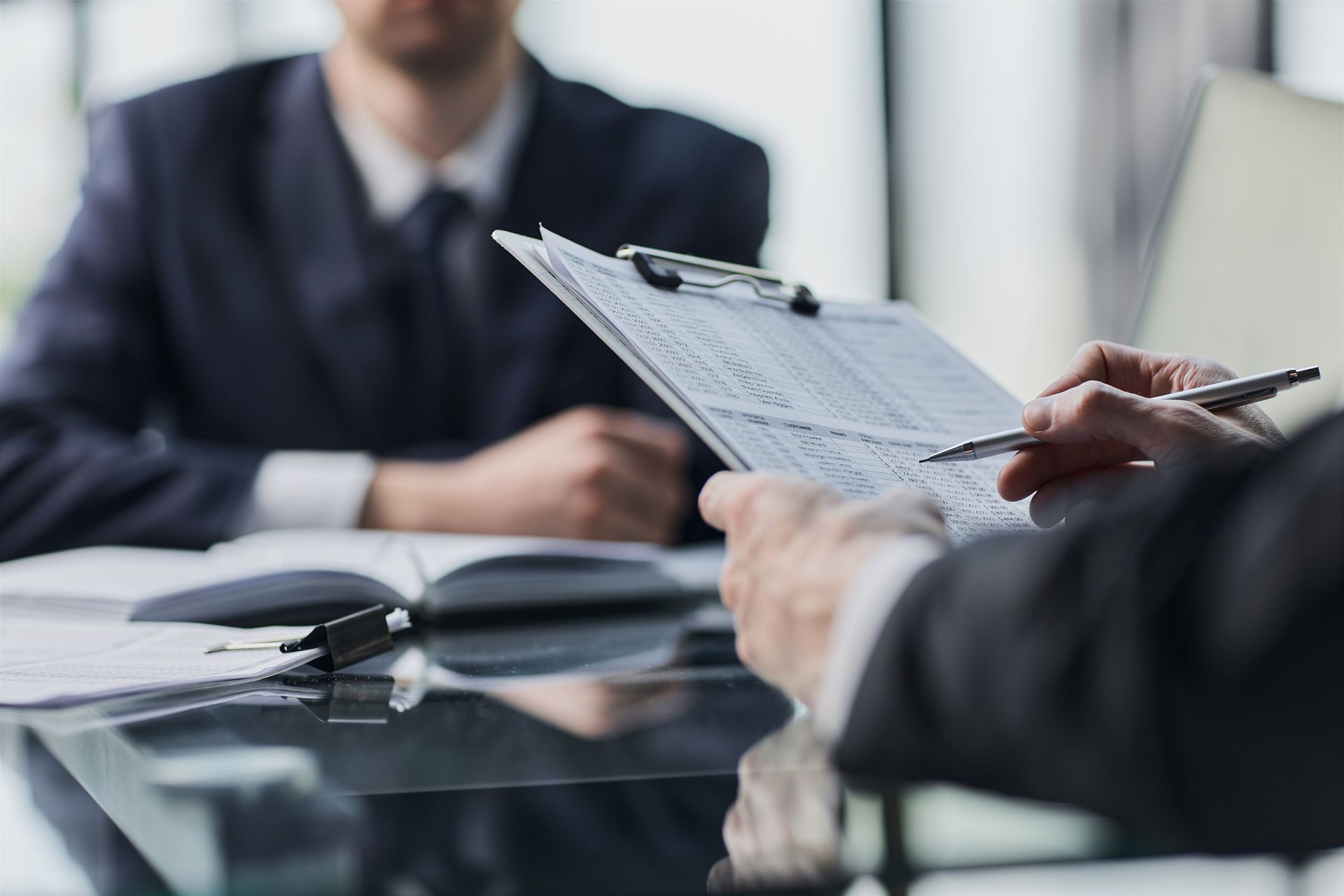 Person taking notes during a meeting at a glass conference table, with a blurred speaker in the background