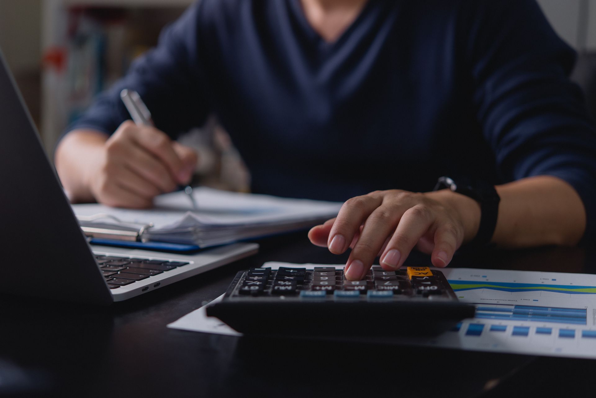 Person using a calculator and writing notes at a desk beside a laptop