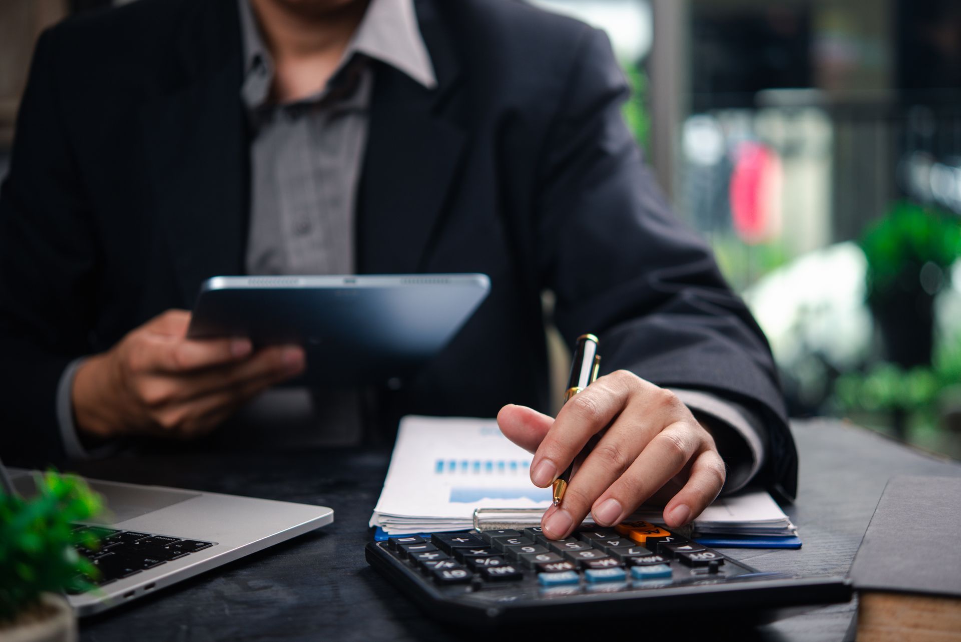 Businessperson using tablet and calculator at a desk with notebook and laptop