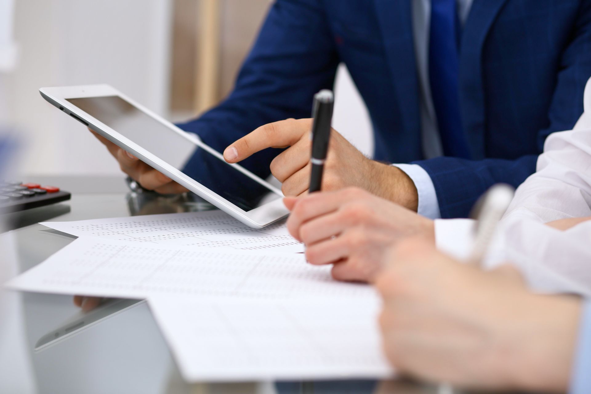People in business attire discuss documents while one uses a tablet and pen at a desk.