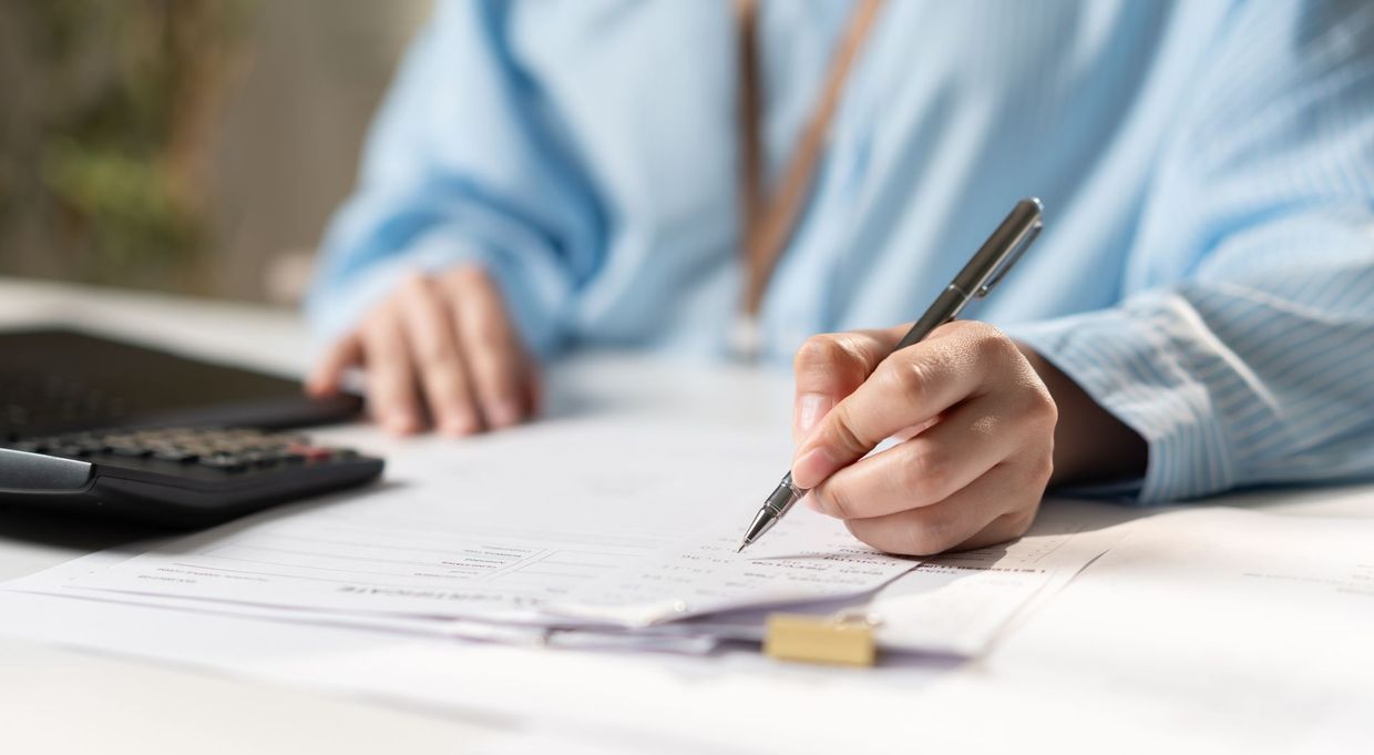 Hand writing on paper beside a calculator on a desk, with a blue shirt sleeve visible