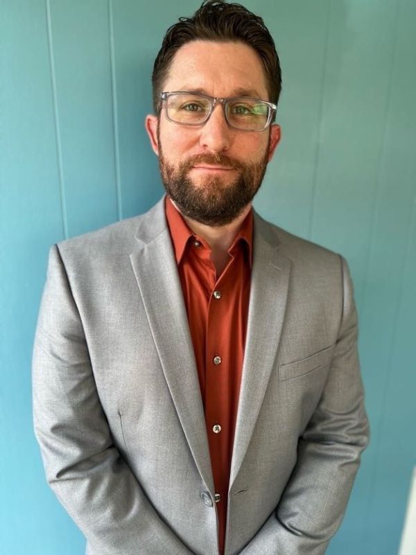 Man in a light gray blazer and rust shirt standing against a teal wall, facing the camera.