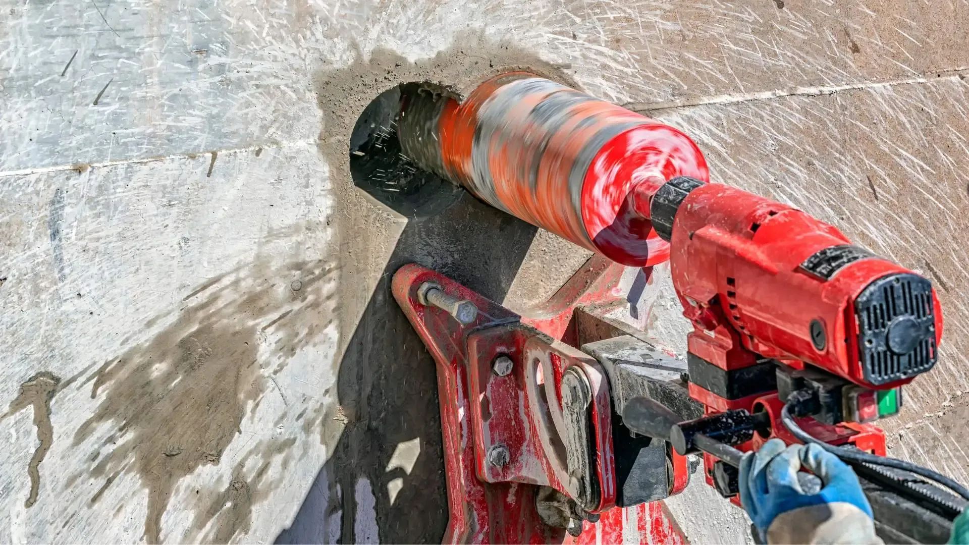 Red core drill boring a hole in concrete. Water present. Person wearing blue glove is operating it.