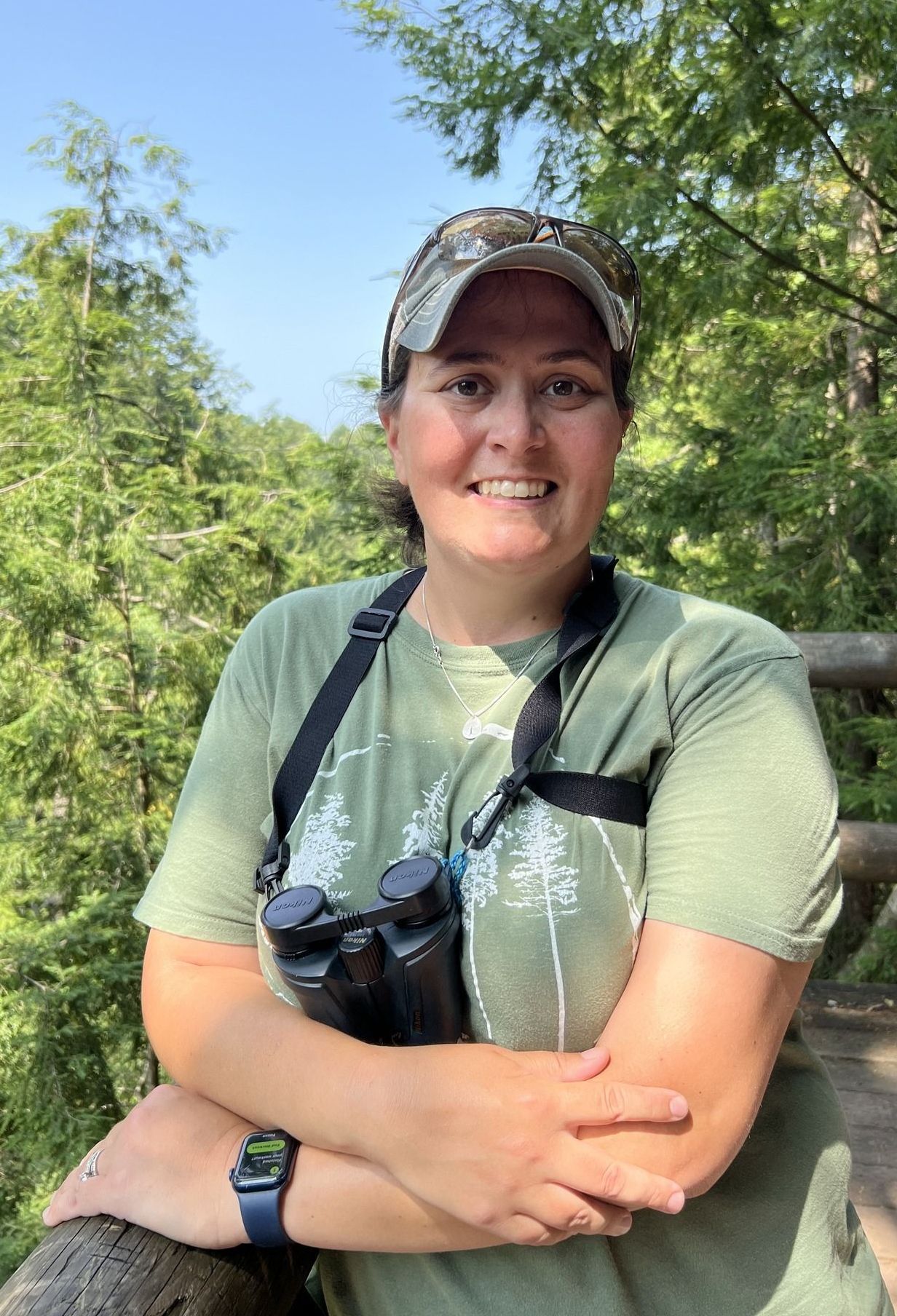 Owner Samantha wearing a green shirt and a hat is holding binoculars while in the mountains.