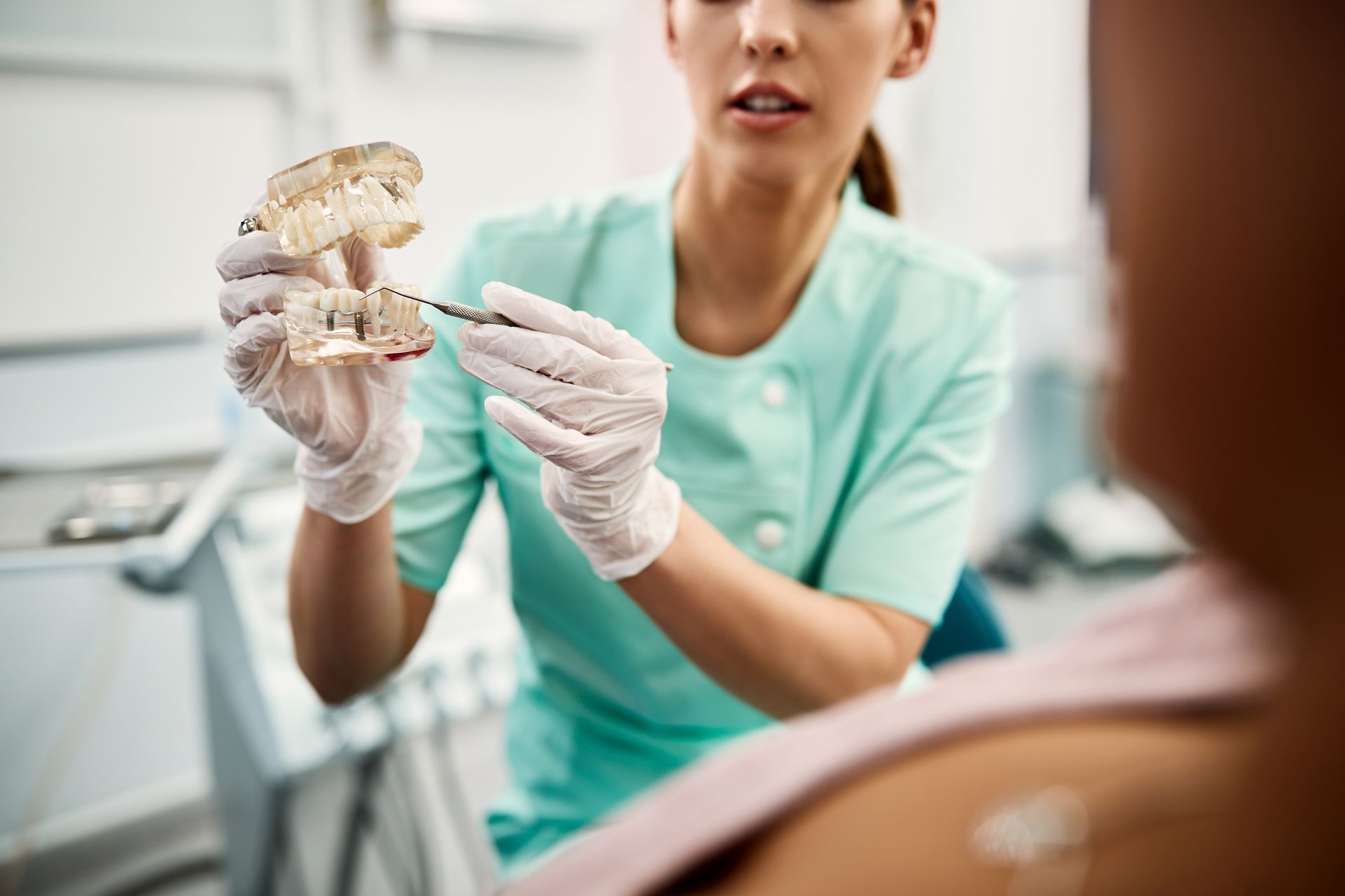 Dentist holding a tooth model during a consultation, wearing teal scrubs and gloves in a clinic room.