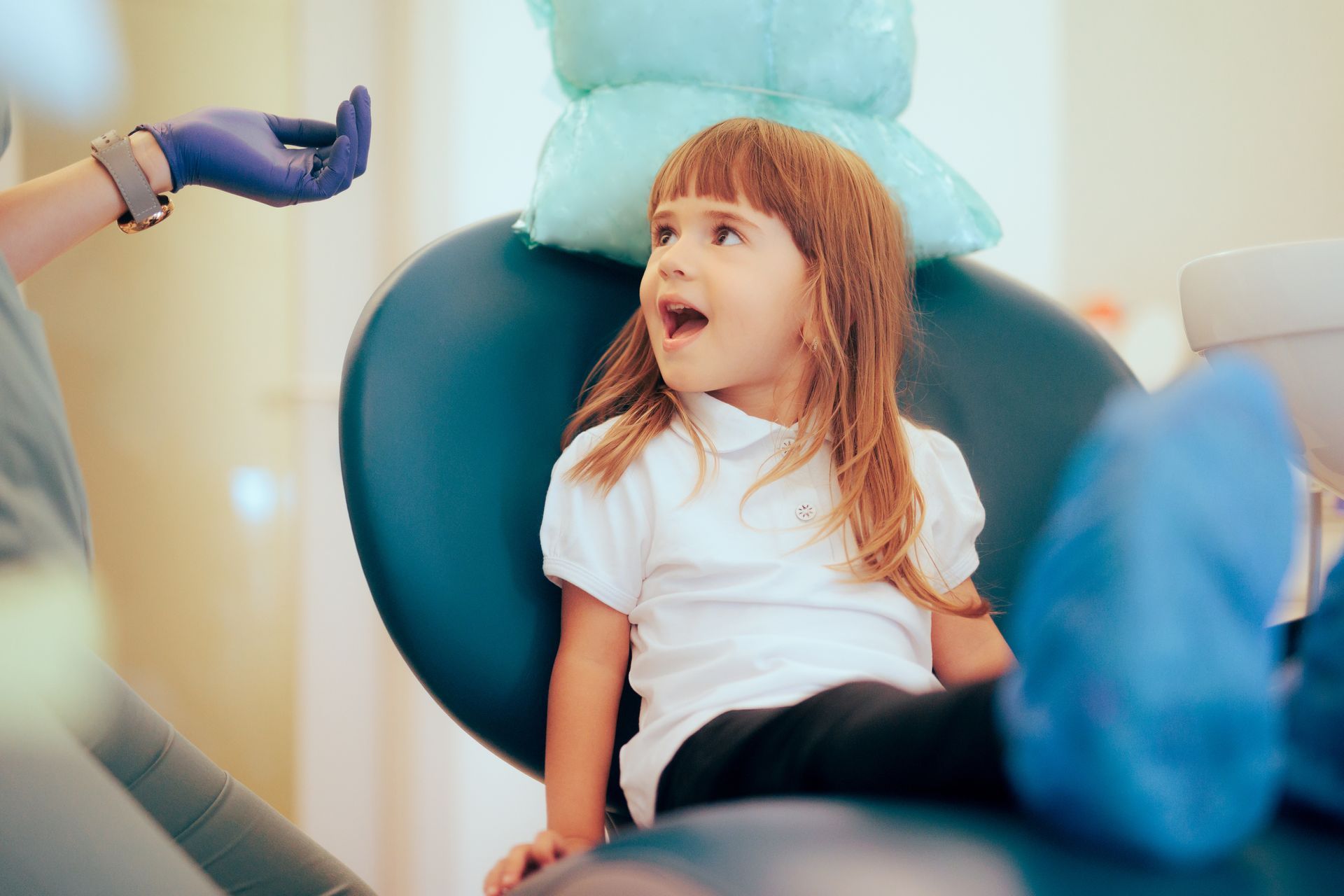 Child in a dentist's chair with an open mouth, while a gloved hand holds a dental tool nearby.