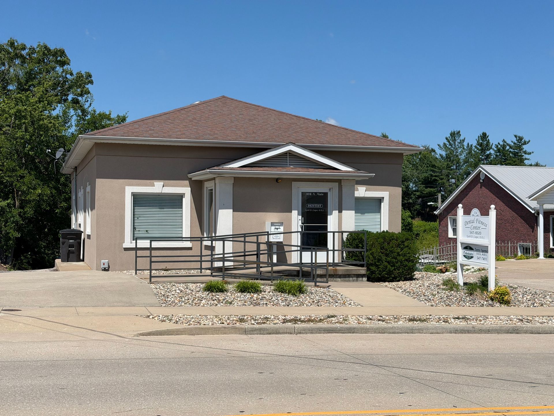 Small beige building with a front porch and ramp, set beside a street under a clear blue sky.