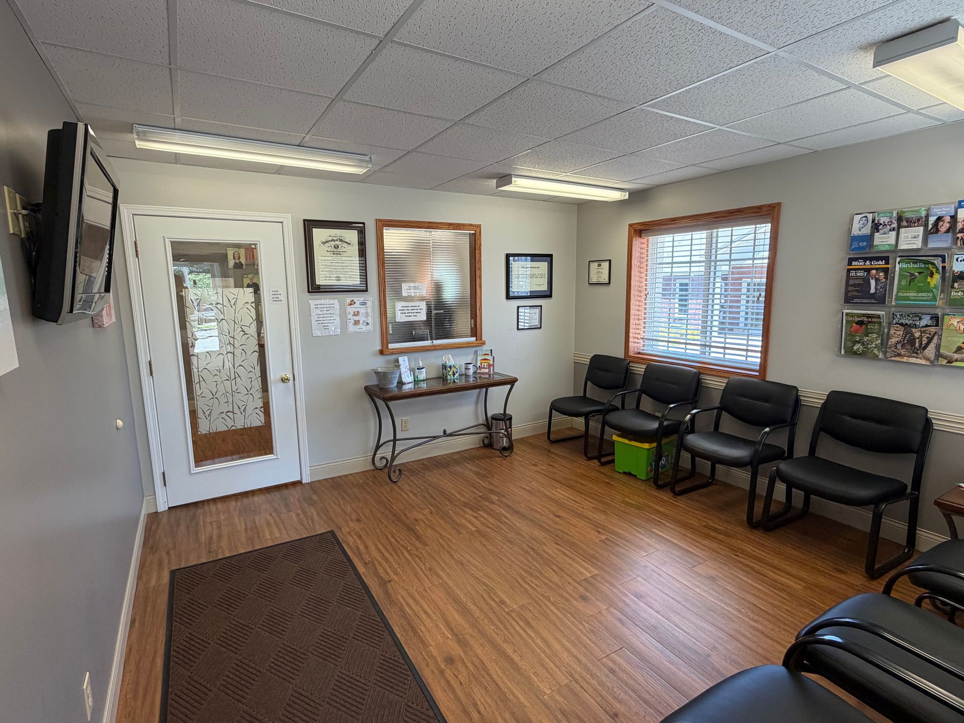 Empty waiting room with black chairs, wood floor, wall art, and a door near the entrance.