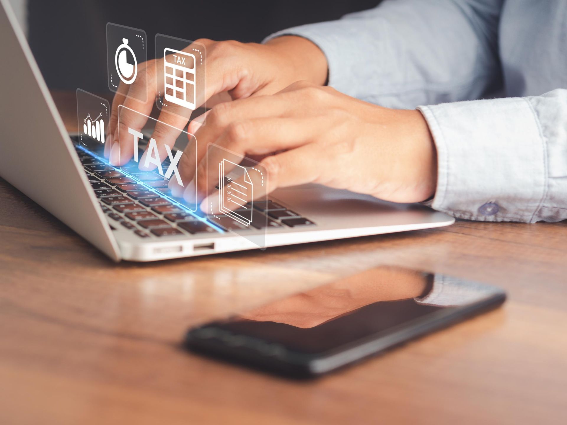Hands typing on a laptop with floating tax icons, next to a smartphone on a wooden desk.