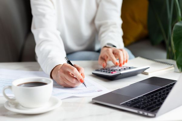 Person writing on paperwork beside a laptop and calculator with a coffee cup on a desk