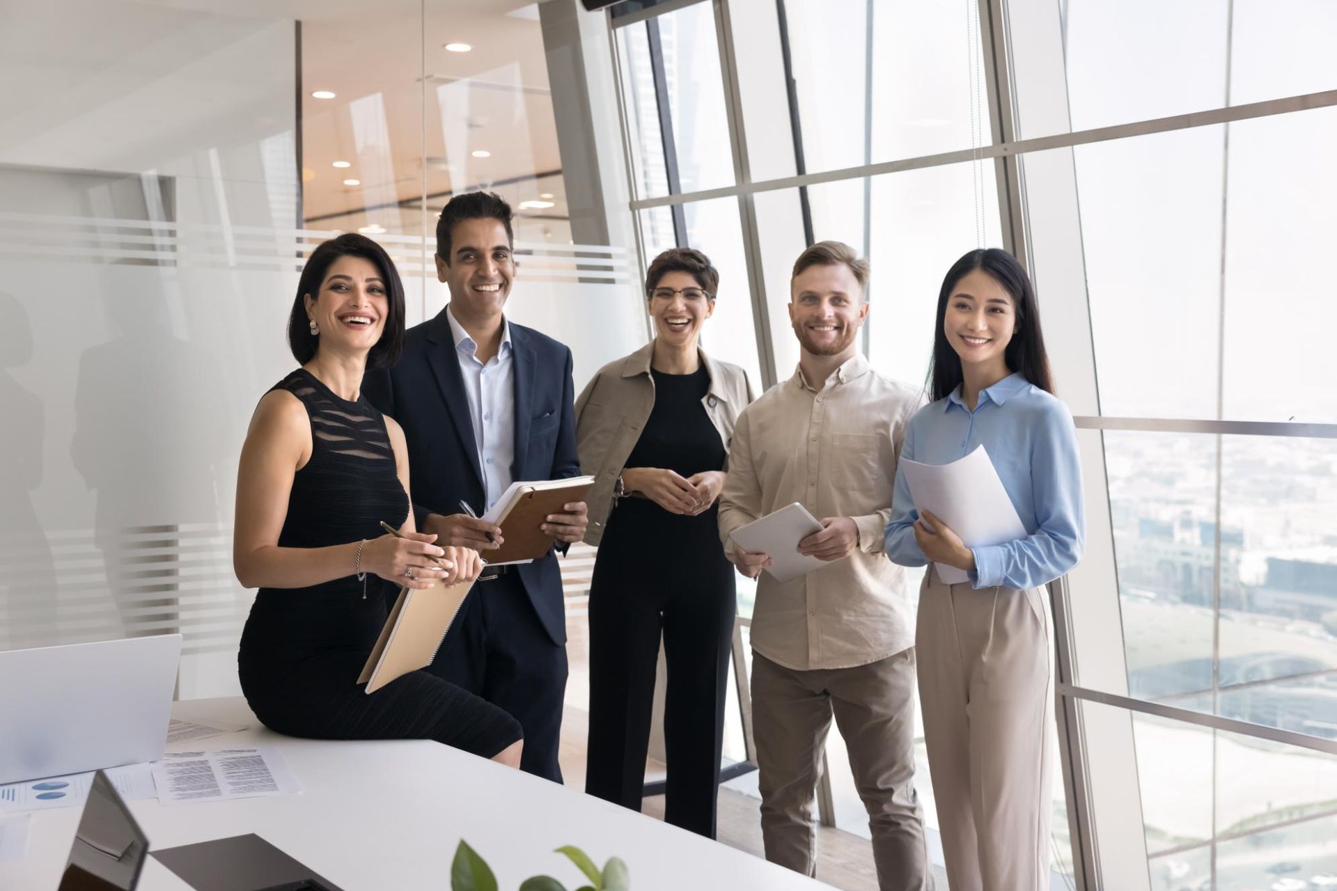 Five coworkers standing in a bright office, smiling and holding tablets and folders.