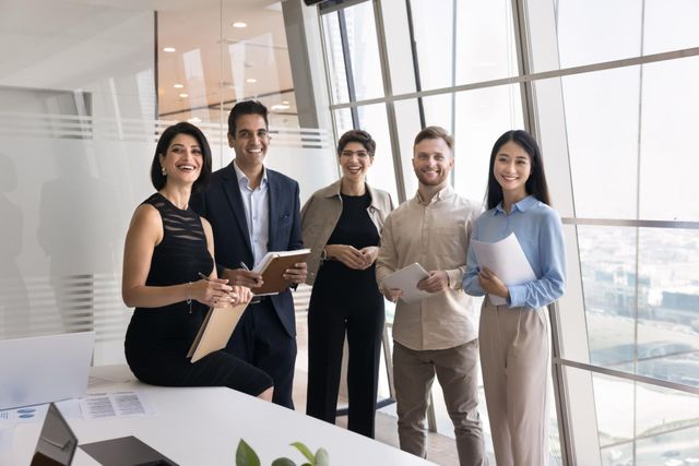 Five coworkers standing in a bright office, smiling and holding tablets and folders.