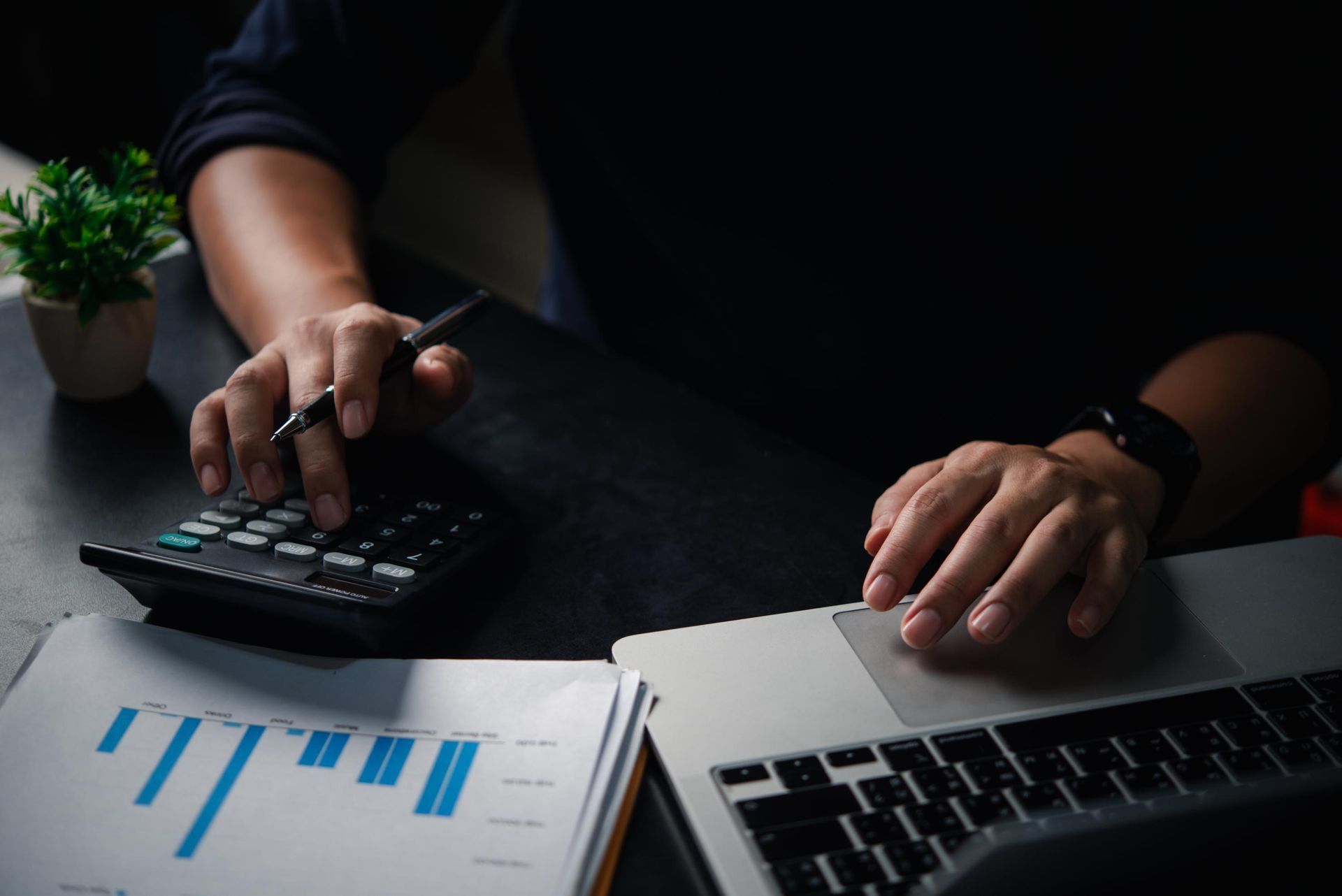 Hands using a calculator and laptop beside a document with charts on a dark desk