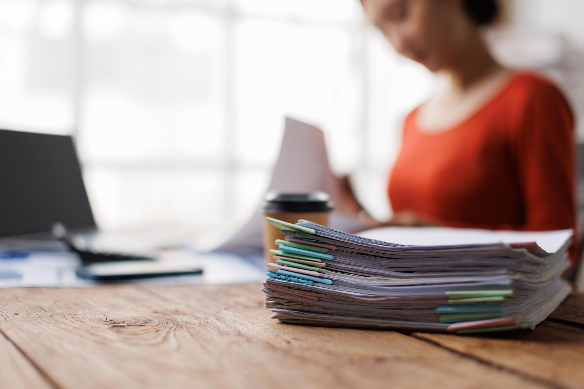 Stack of papers on a wooden desk with a blurred person in a red top working in the background
