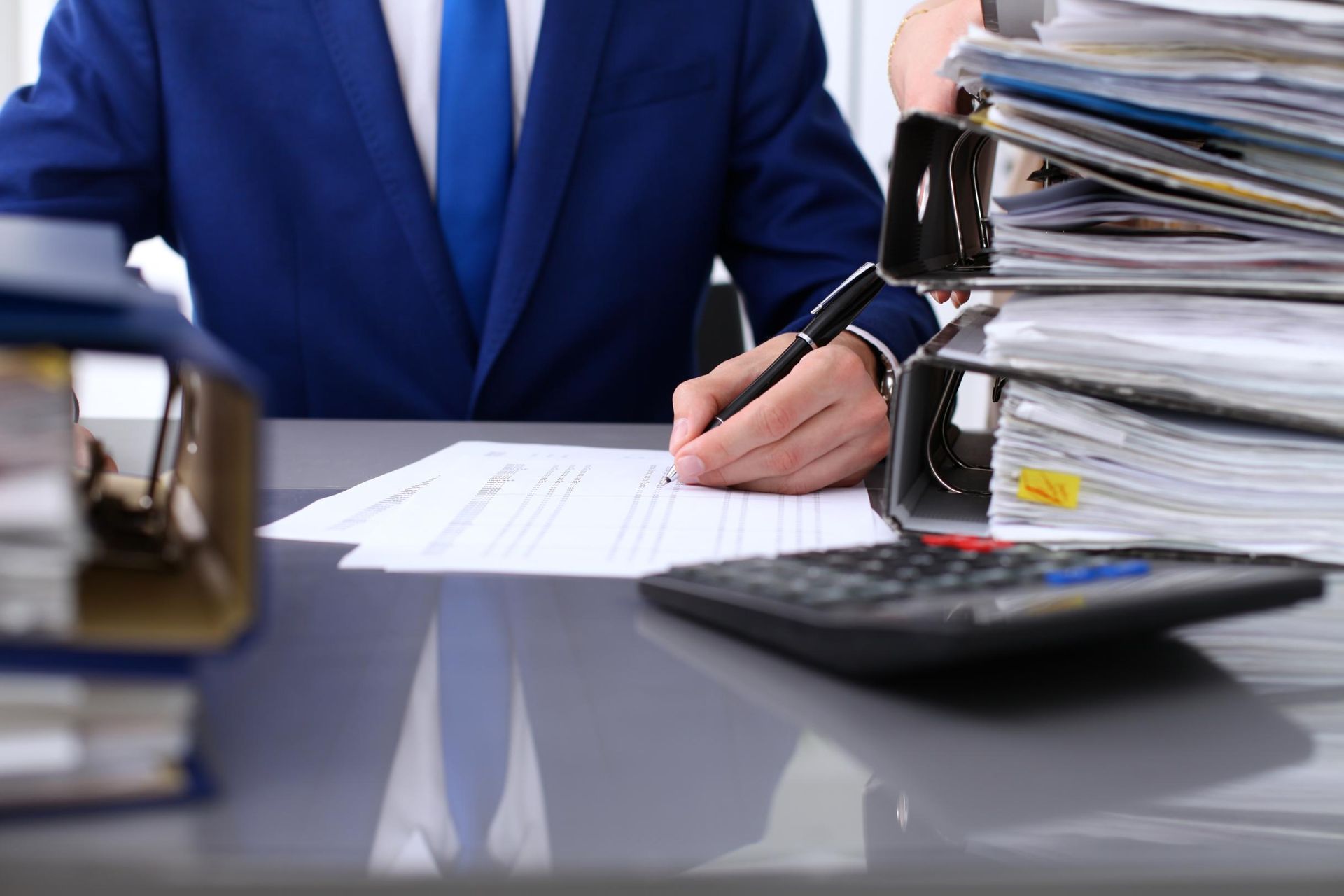 Person in a blue suit writing at a desk with stacks of papers and a calculator nearby