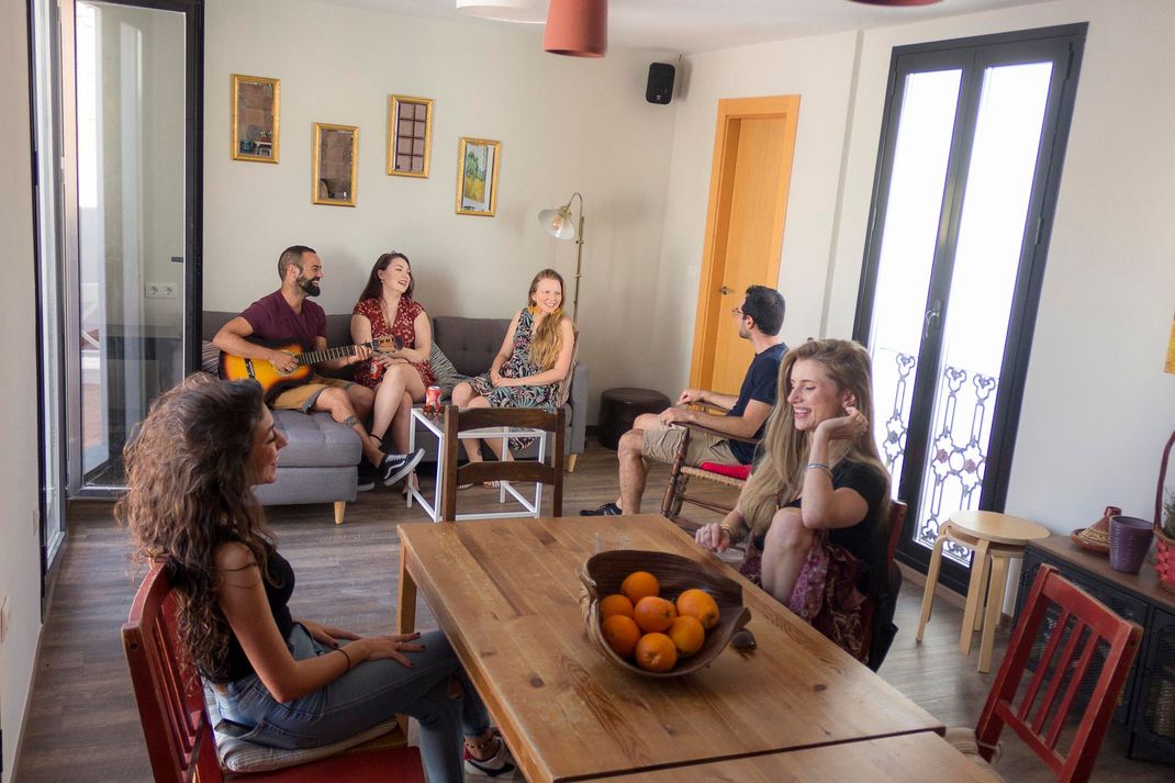 People seated and chatting in a bright living room around a wooden table with a bowl of oranges.