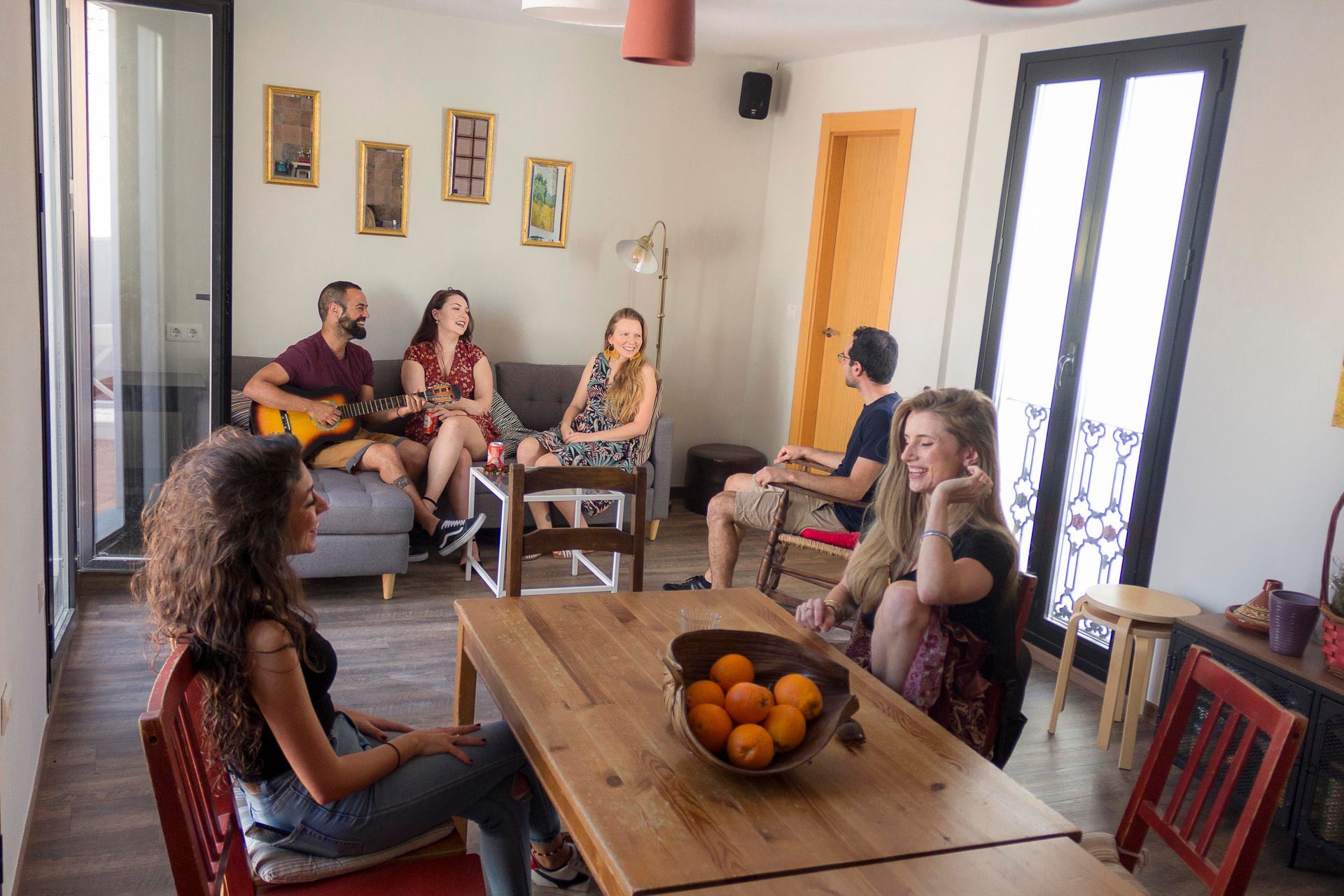 People seated and chatting in a bright living room around a wooden table with a bowl of oranges.
