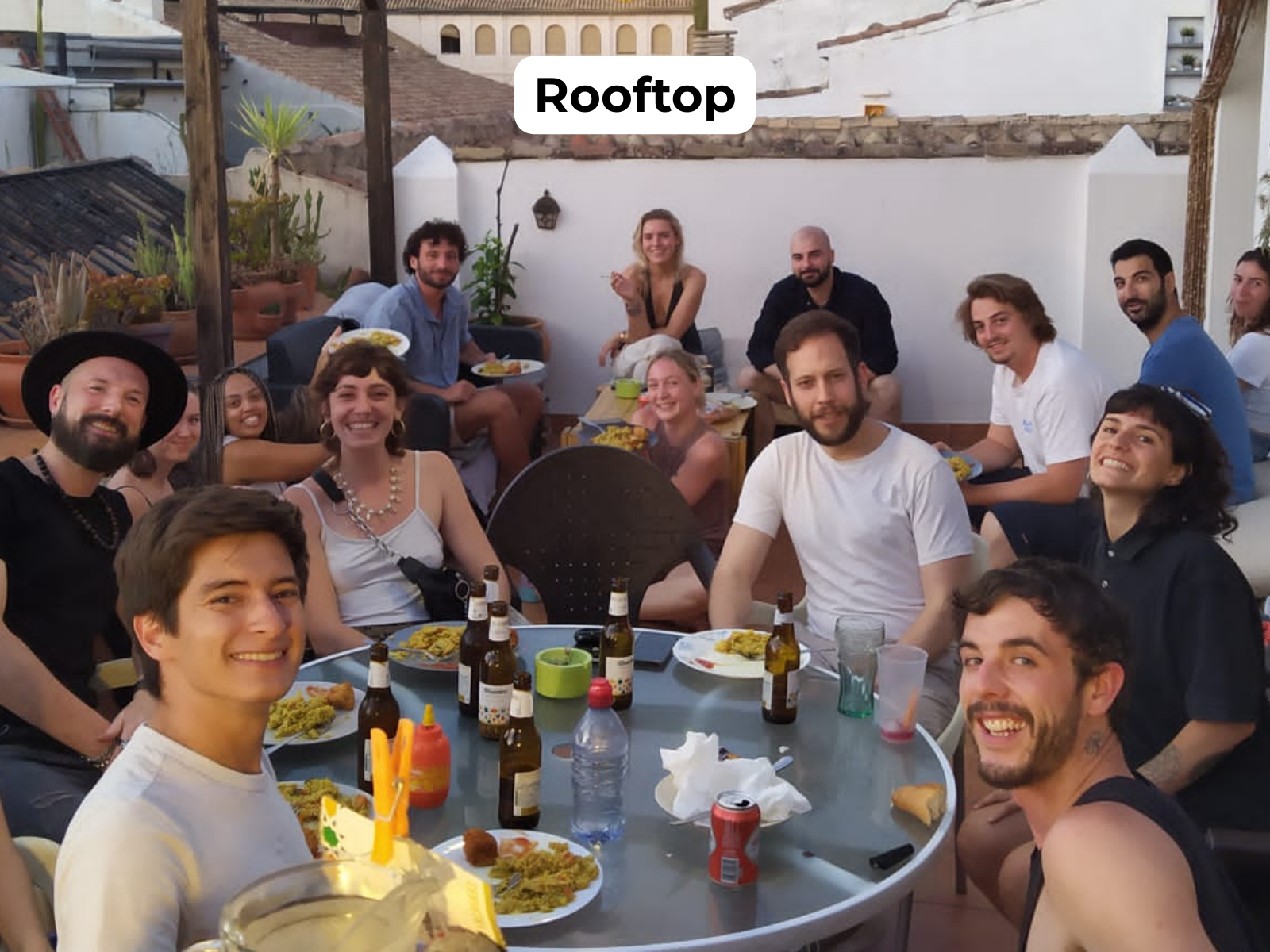 Group of people smiling around a table on a rooftop patio, with drinks and food in warm evening light.