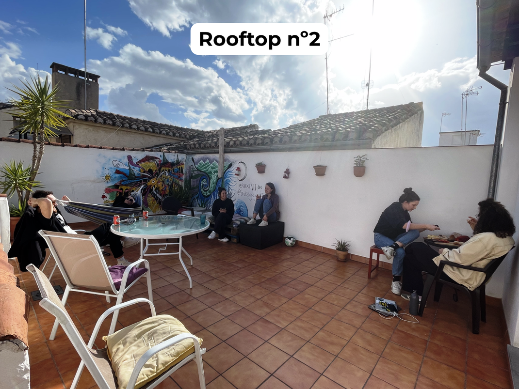 People relaxing on a rooftop terrace with white walls, terracotta tiles, and sunny sky.