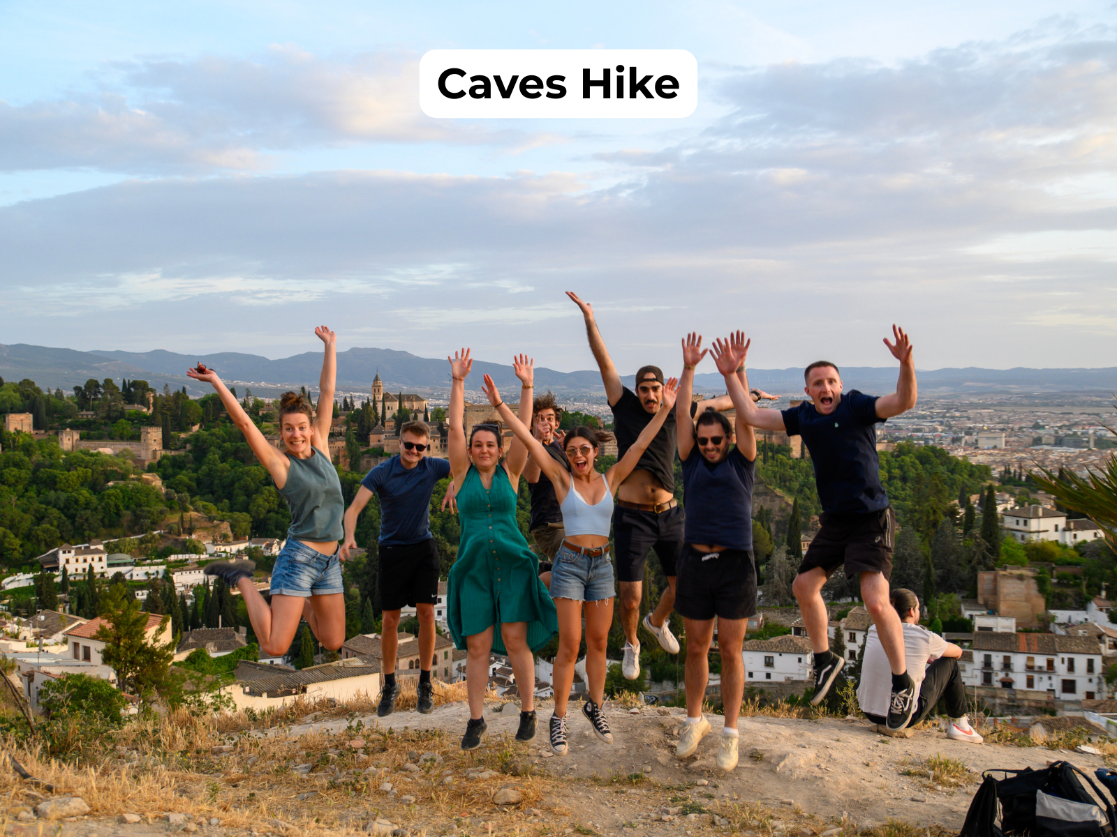 Group of hikers jumping with arms raised on a rocky hill, city skyline in the background, titled “Caves Hike”