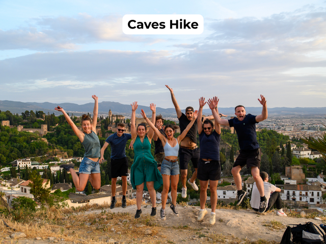 Group of hikers jumping with arms raised on a rocky hill, city skyline in the background, titled “Caves Hike”