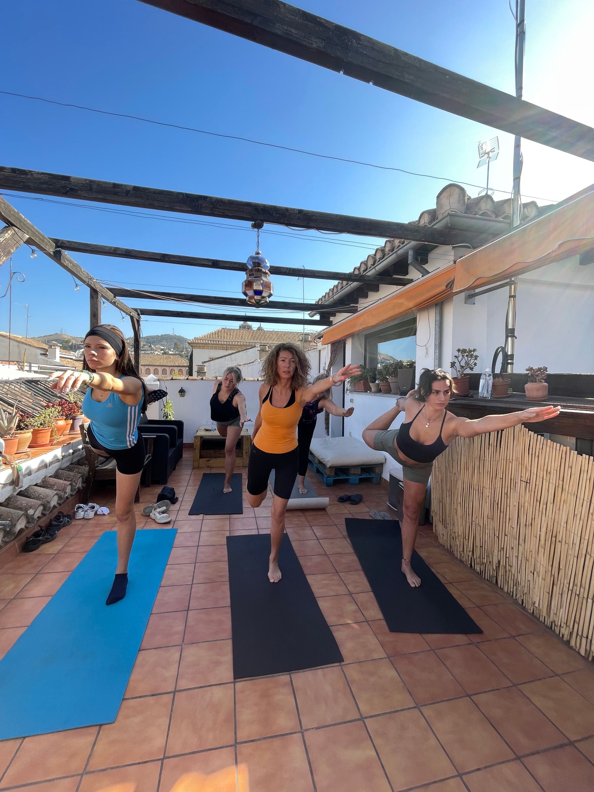 People doing yoga on mats outdoors on a sunny rooftop patio with blue sky and pergola beams