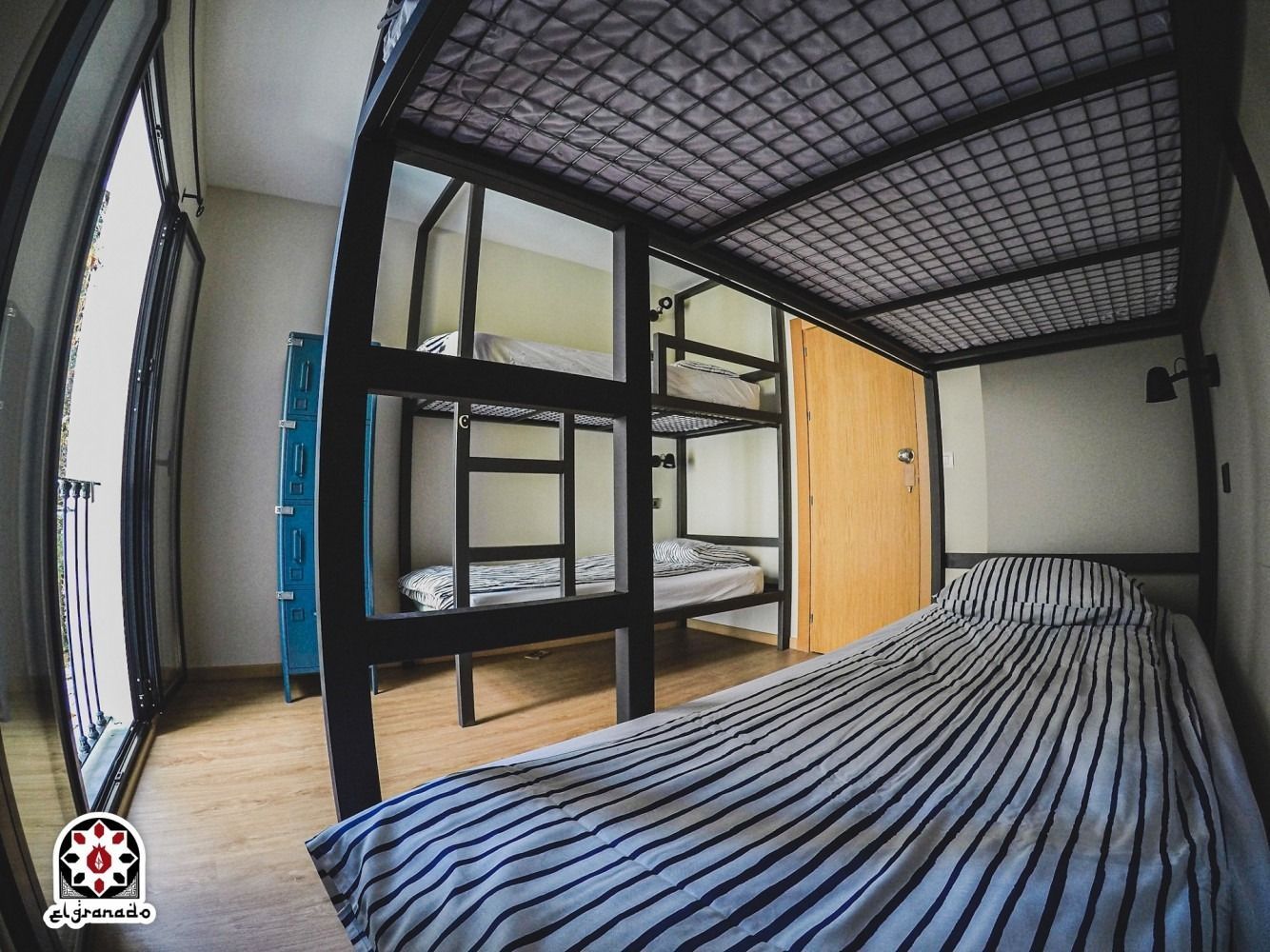 Dorm room with bunk bed, striped bedding, wooden door, and lockers by a large window.