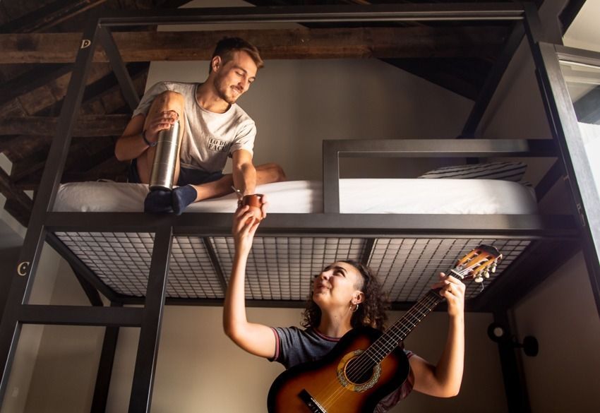 Two people on a bunk bed, reaching down and up toward each other, one holding a guitar.