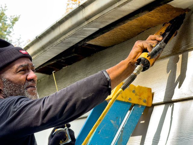 Man uses caulking gun on a house exterior, standing on a ladder.