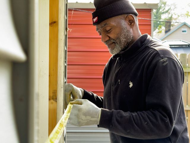 Man in black shirt and beanie measures a wooden structure with a tape measure outdoors.