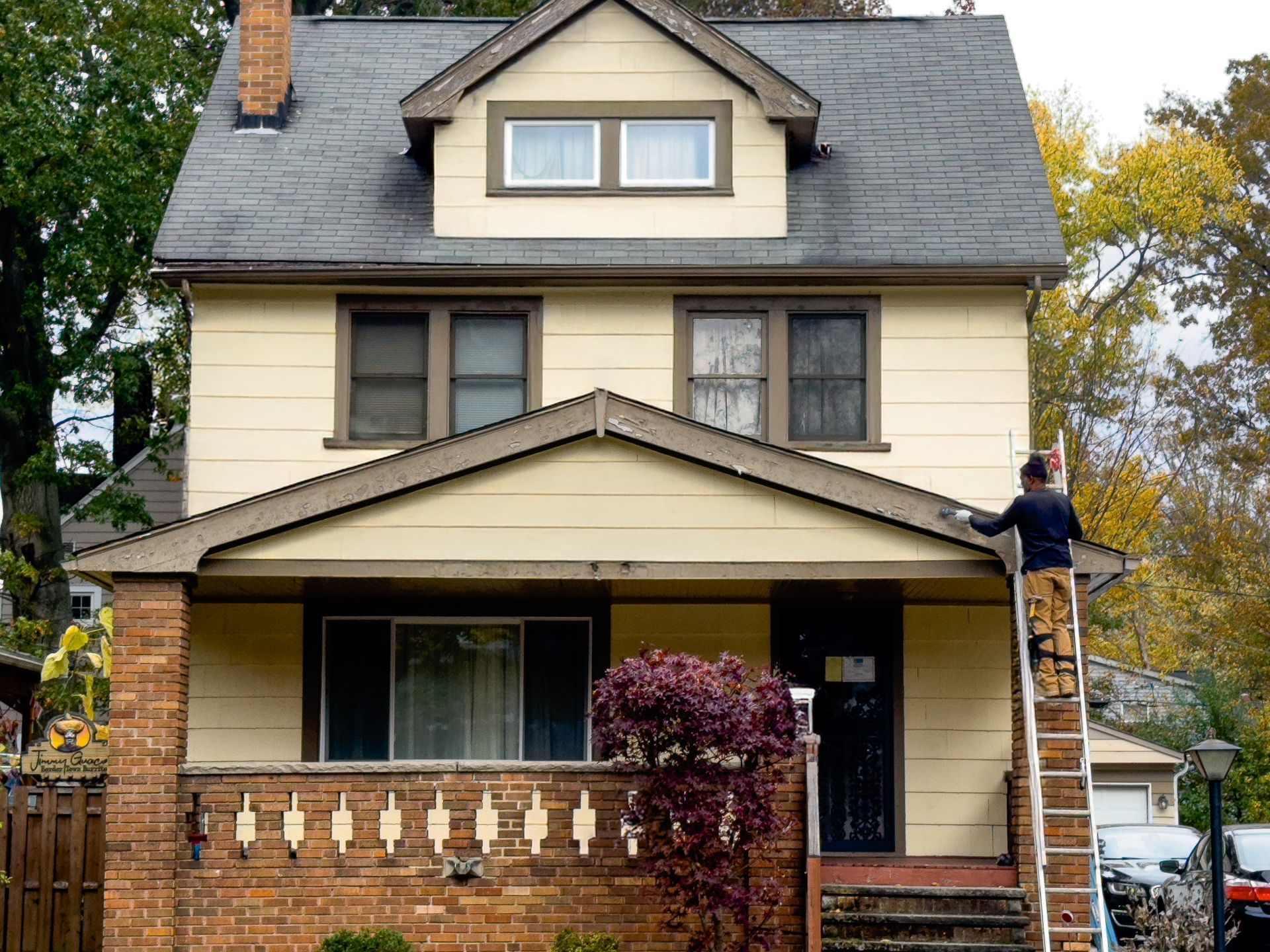 Person on a ladder painting the trim of a two-story beige house with a brick base.