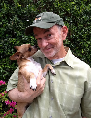 Man in green cap holding a small dog, both beside a leafy hedge outdoors.