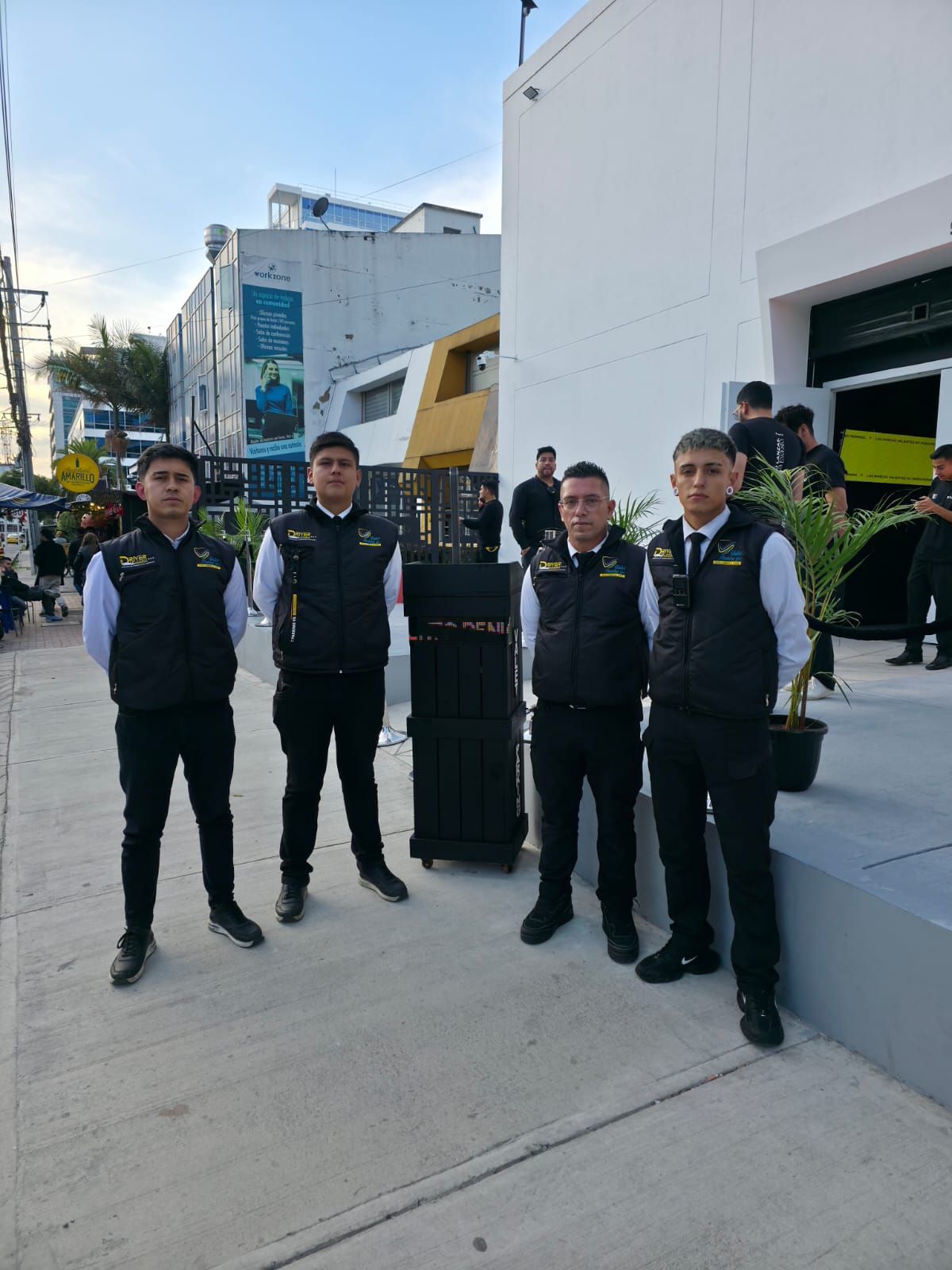 Cinco guardias de seguridad uniformados permanecen de pie en una acera junto a un edificio blanco y una barandilla negra.