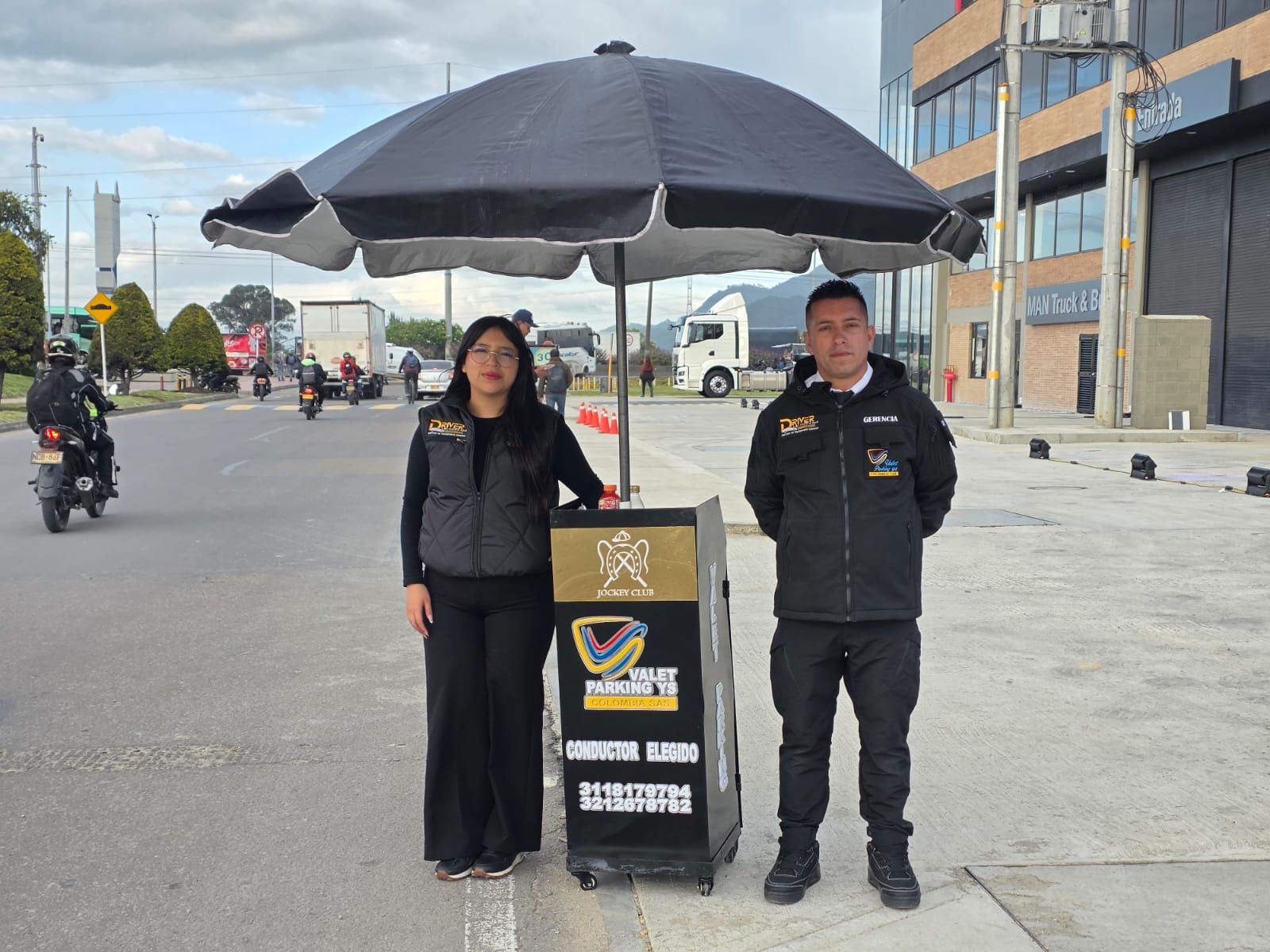 Dos personas de pie bajo una gran sombrilla negra junto a una calle, con un cartel delante de ellas.