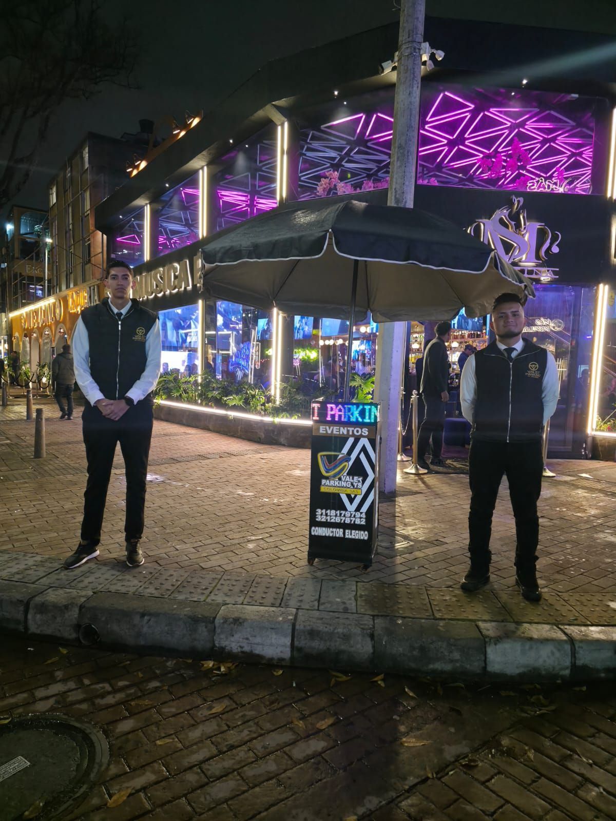 Dos personas de pie frente a un restaurante iluminado con luces de neón por la noche, con paraguas y un pavimento empedrado.