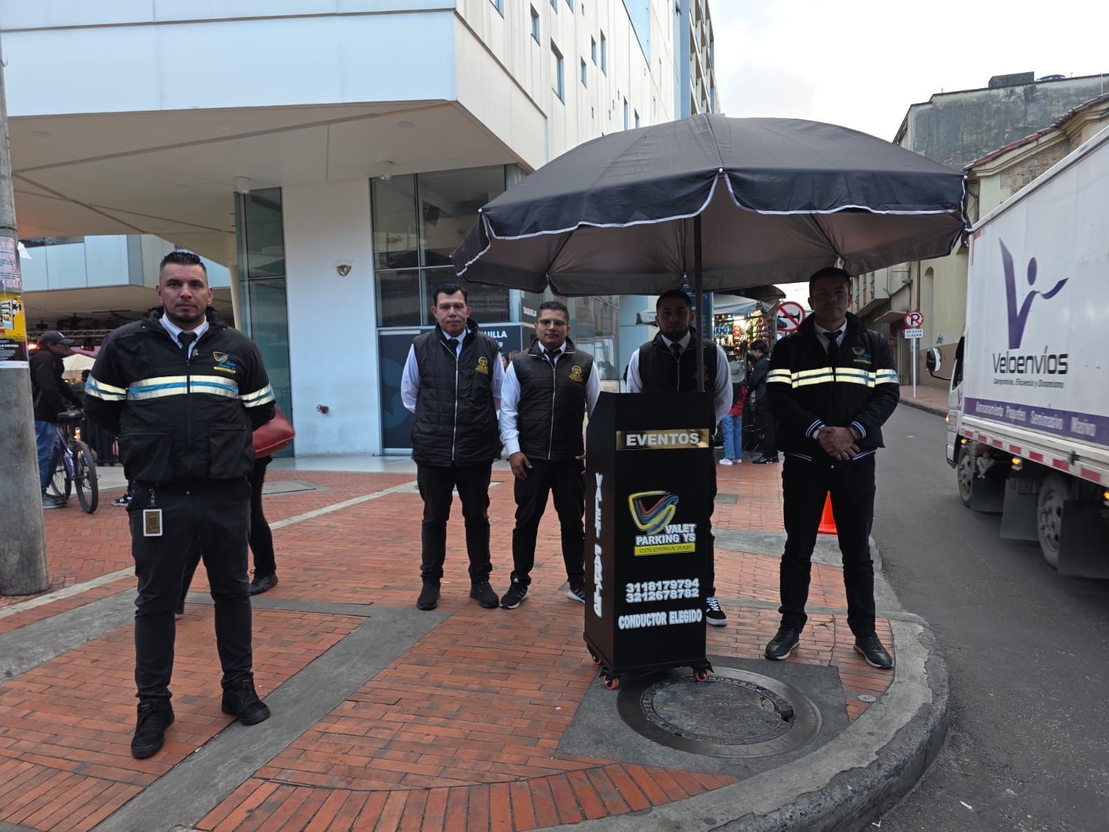 Personal de seguridad uniformado de pie junto a un paraguas negro y una barricada en una calle adoquinada cerca de un camión.