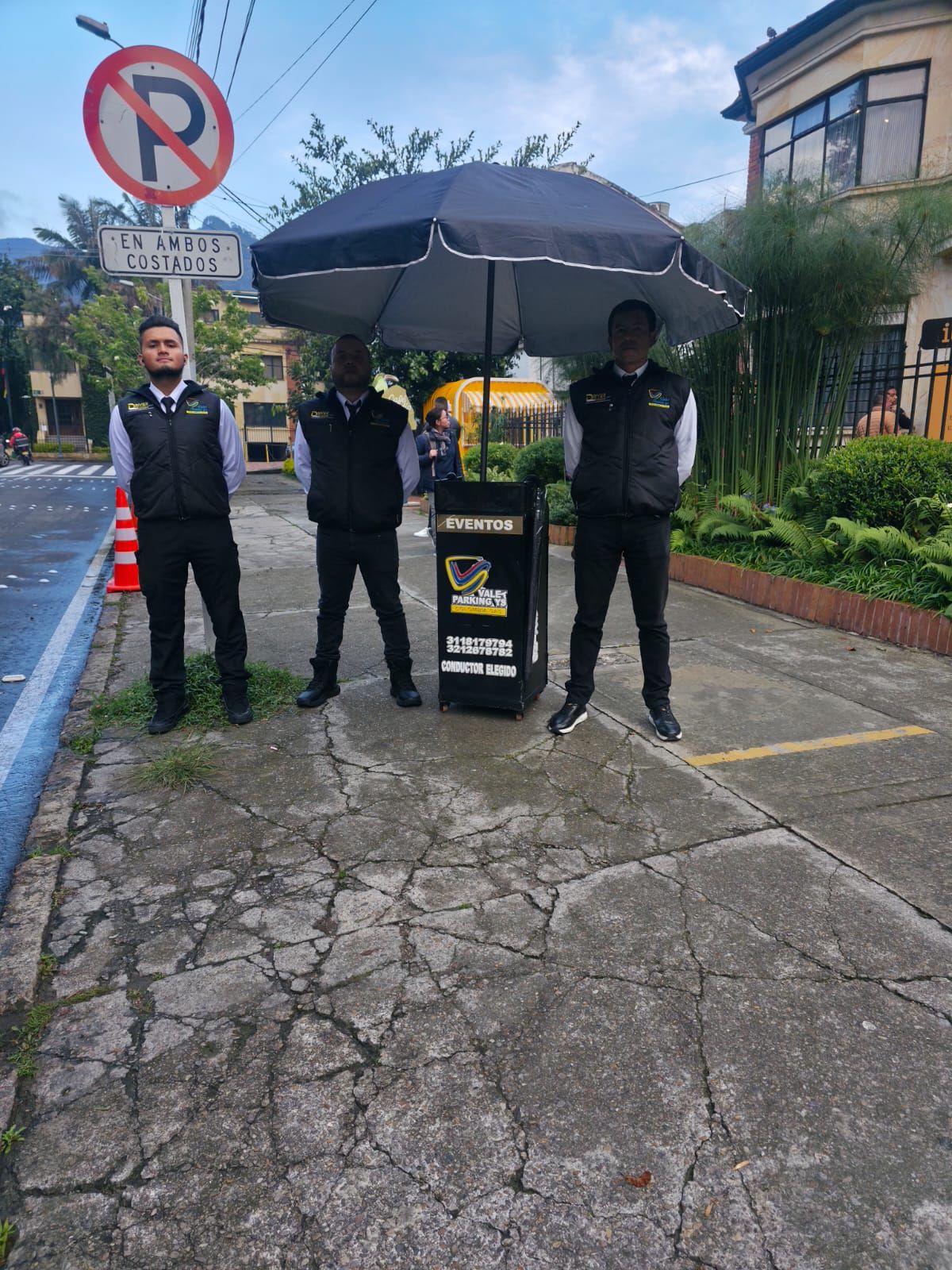 Tres hombres uniformados permanecen de pie bajo un paraguas junto a un letrero y un podio en una calle mojada.