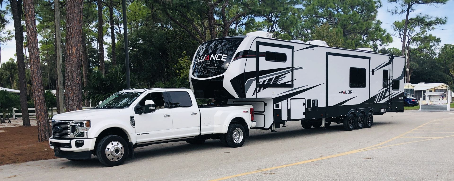 White pickup truck towing a large black-and-white fifth-wheel RV on a roadside near trees