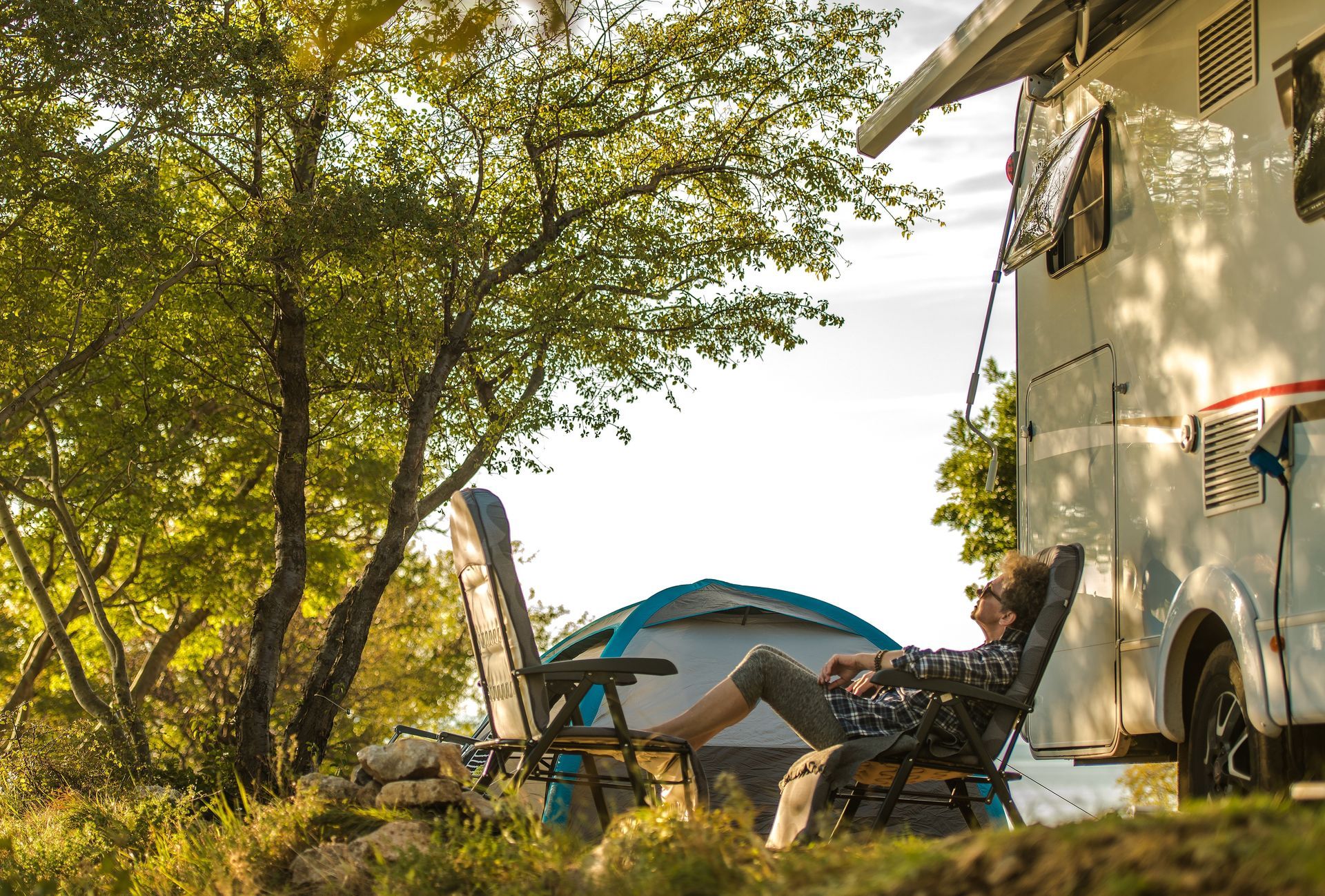 Camp chairs beside a camper under trees, with a small tent in the background at a sunny campsite.