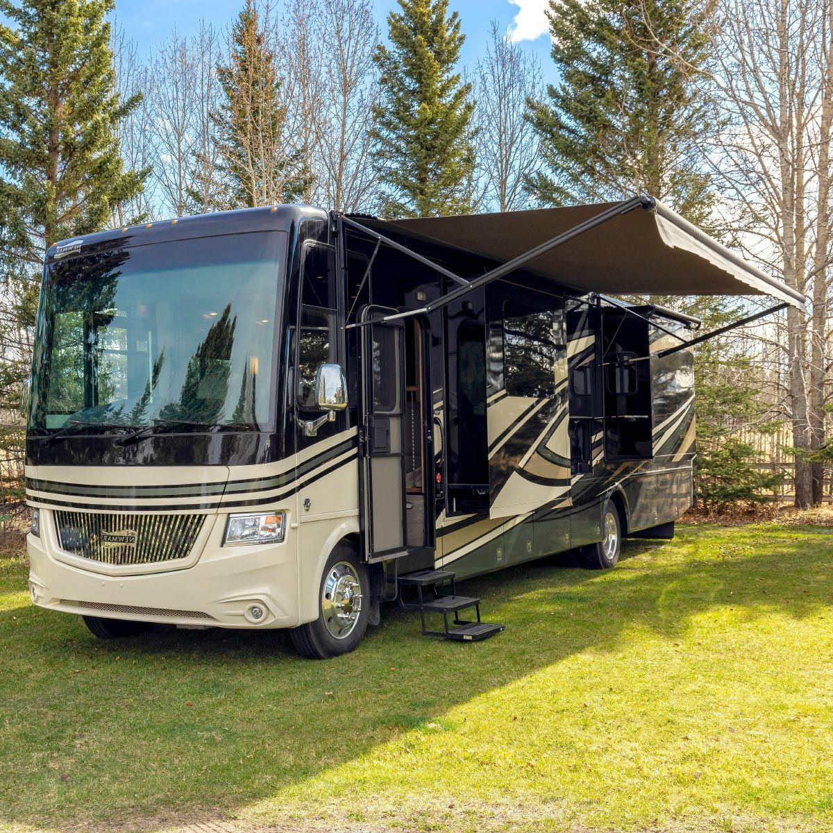Large beige motorhome with extended awning parked on a grassy lawn among trees