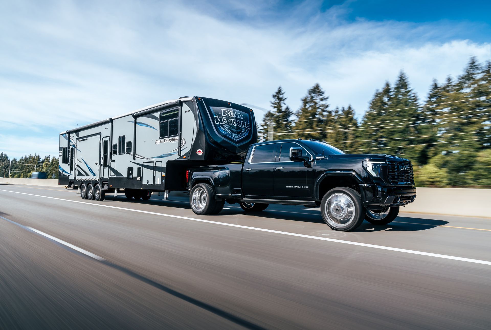 Black pickup truck towing a large white RV trailer on a highway with trees in the background